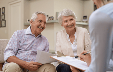 A happy couple smiling and discussing with a real estate agent, seated together at a table, with documents and a property listing in front of them, conveying a sense of excitement and collaboration.