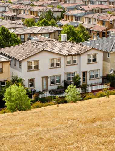 A view of a suburban neighborhood with many houses, most with brown tiled roofs, nestled on a rolling hill.